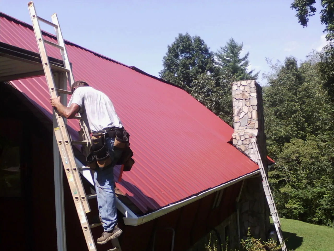 Roofer on a ladder beside a red metal roof and stone chimney — Soffit & Fascia Repair by Roofing Brothers in Shirleysburg